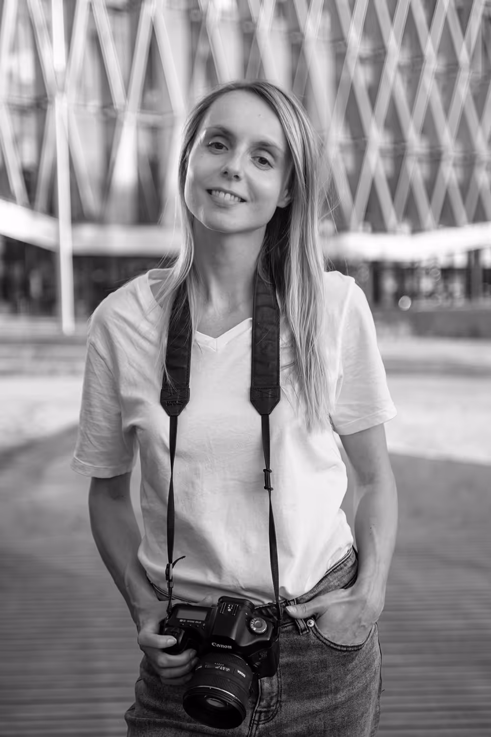 Black and white lifestyle portrait of photographer Kasia Bryjak, smiling and standing with a Canon camera hanging from her neck, set against a modern architectural background with a symmetrical, blurred pattern.